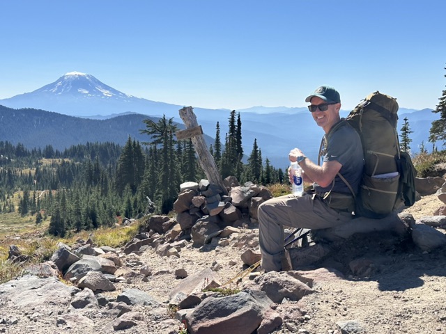 Jesse resting on a rocky trail with Mount Adams in the distance